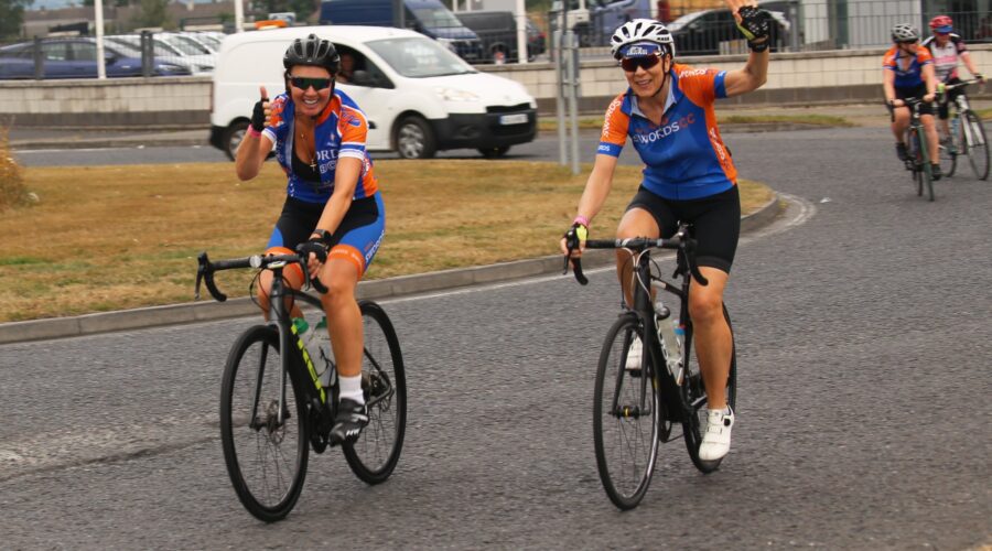 Two women wave to camera while cycling bikes.