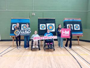 Group of girls hold signage for Her Moves while next to archery targets.