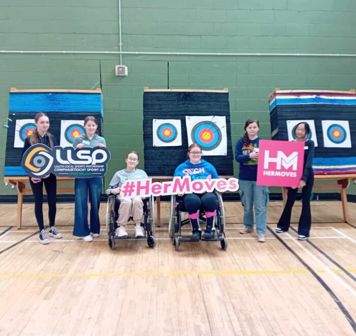 Group of girls hold signage for Her Moves while next to archery targets.