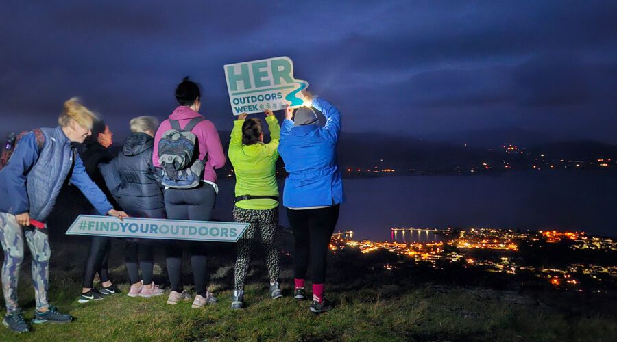Women hold Her Outdoors signage with backs turned and facing the night sky of Cooley's landscape.