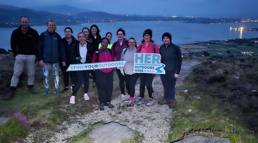 Group of people hold signage for Her Outdoors Week with Cooley landscape in background.