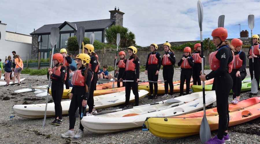Young girls hold oars while wearing wetsuits and life vests.