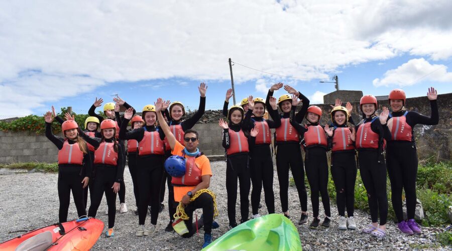 Young girls in wetsuits and life vests wave towards camera.
