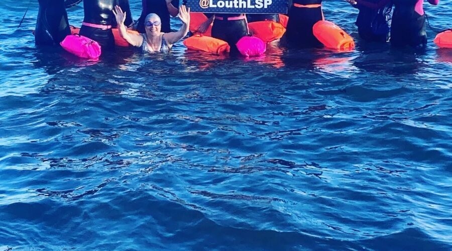 Women in ocean wave and hold sign for Louth LSP.