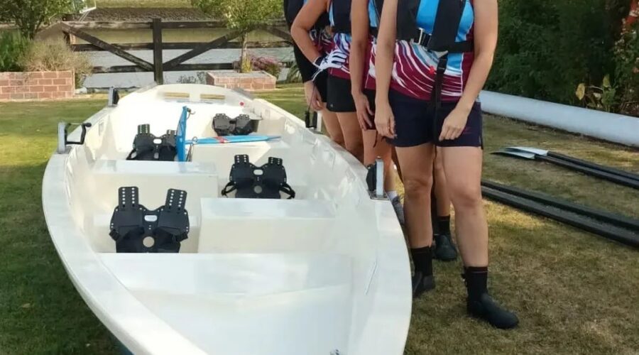 Four women stand by river rowing boat.