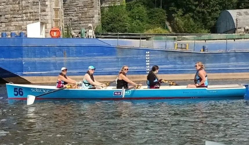 Five women pictured on a rowing boat, rowing beneath a bridge.