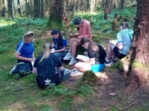 Girl sits on tree stump while others crouch down to draw in notebooks.