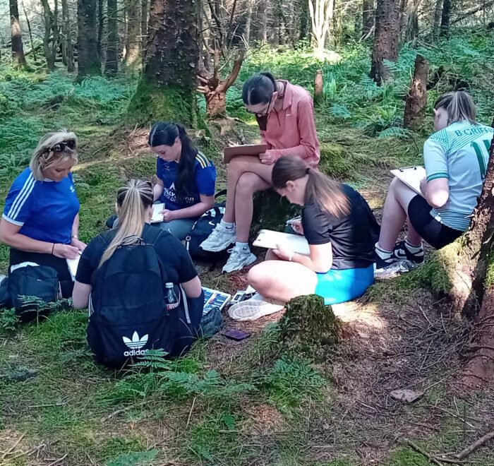 Girl sits on tree stump while others crouch down to draw in notebooks.