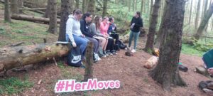 Girls draw on a clipboard while sitting on fallen tree trunk.