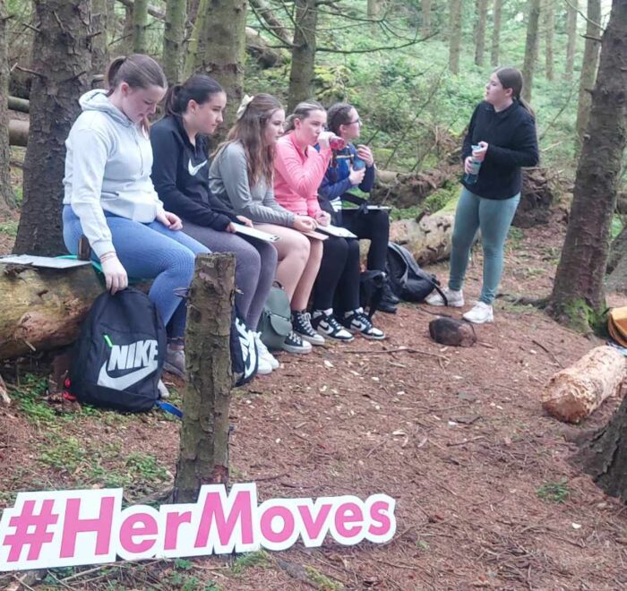 Girls draw on a clipboard while sitting on fallen tree trunk.