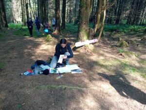 Young girl kneels on ground in forest and checks paper.