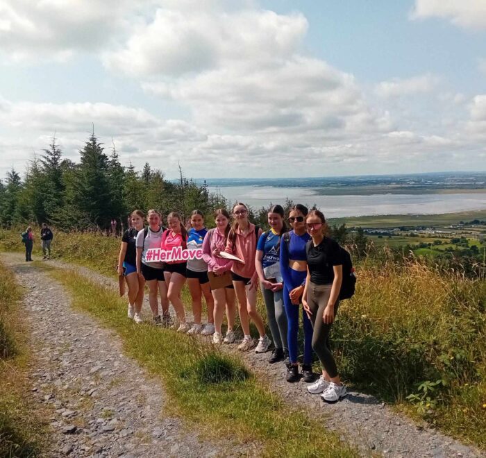 Group of girls hold sign for Her Moves along forest path.