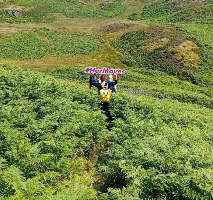 Girl hold up sign for Her Moves above head with back turned to camera in a field of green grass.