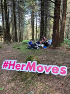 Group of girls stand in circle near trees in forest with sign for Her Moves in forefront.