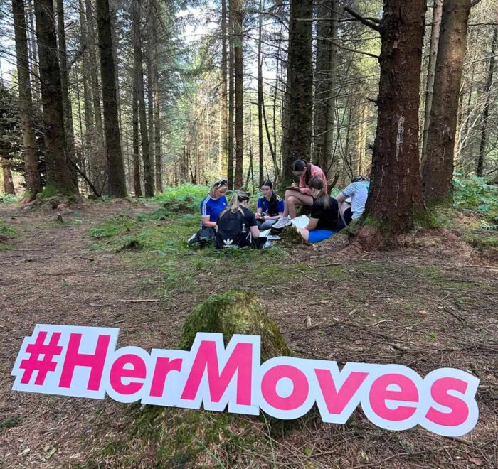 Group of girls stand in circle near trees in forest with sign for Her Moves in forefront.