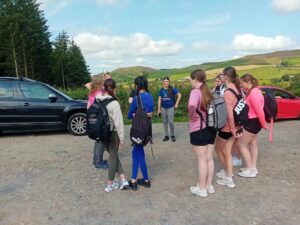 Group of girls in gym gear and school bags stand in a circle near forest.