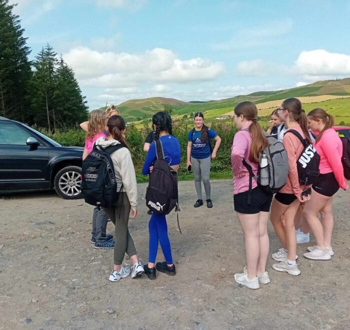 Group of girls in gym gear and school bags stand in a circle near forest.