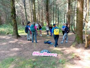 Group of girls stand in circle near trees in forest with sign for Her Moves in forefront.