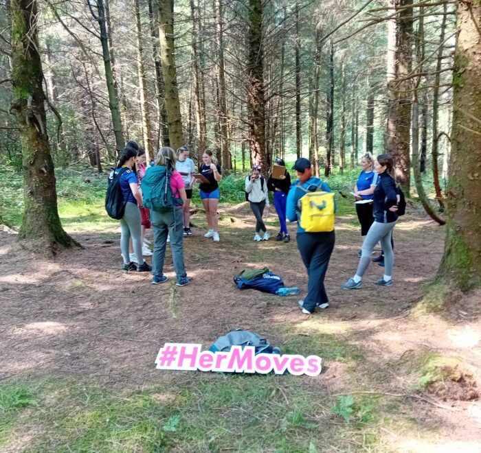 Group of girls stand in circle near trees in forest with sign for Her Moves in forefront.