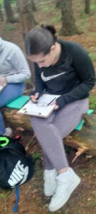 Girl draws on a clipboard while sitting on fallen tree trunk.