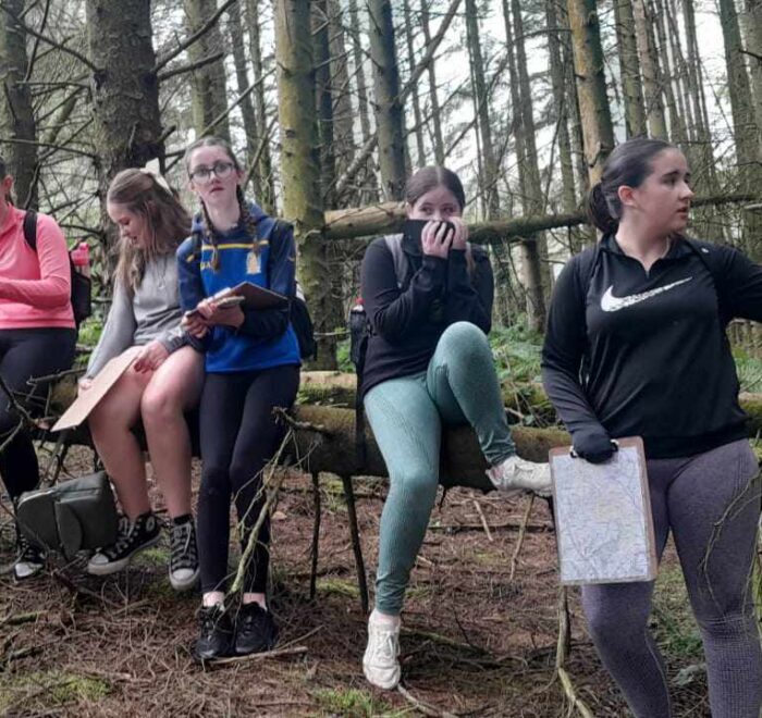 Group of girls sit on fallen tree trunk in forest.
