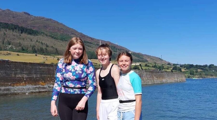 Three young girls stand at water's edge.
