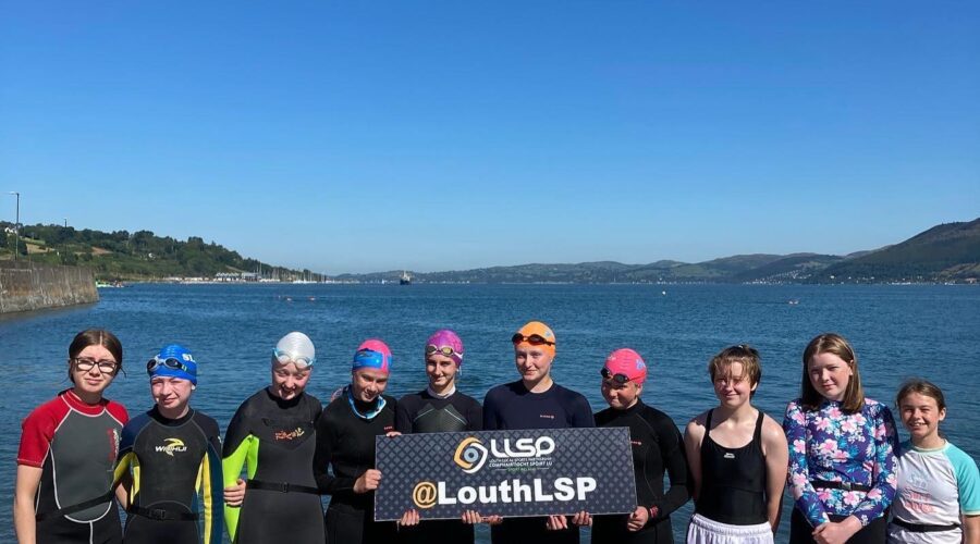 Women in wetsuits hold signage for Louth LSP at waters edge.