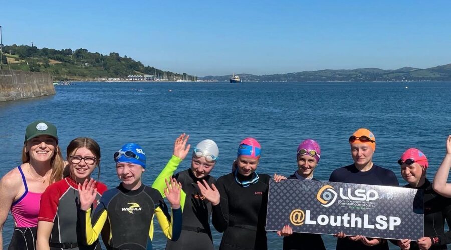Women in wetsuits hold Louth LSP sign at the sea's edge.
