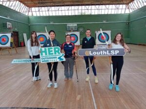 Five girls hold archery gear in gym.