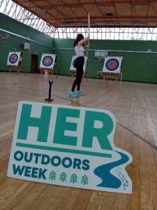 Teenage girl plays archery with back turned to camera and sign for Her Outdoors is in the forefront.