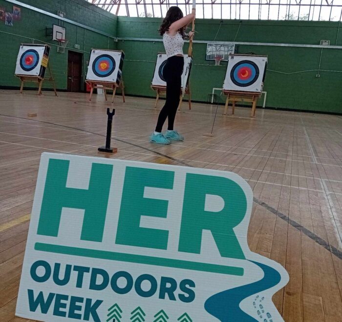 Teenage girl plays archery with back turned to camera and sign for Her Outdoors is in the forefront.
