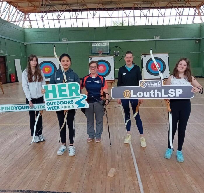 Five girls hold archery gear in gym.