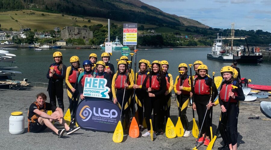 Young girls pose near water in wetsuits and life vests while holding oars.