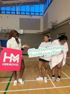 Group of girls hold signage for Her Moves in gym.