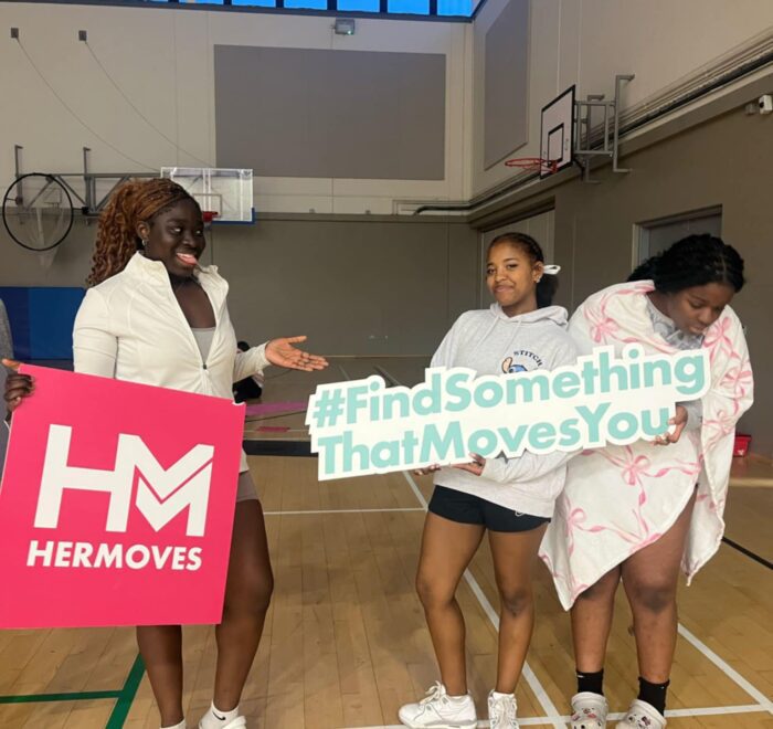 Group of girls hold signage for Her Moves in gym.