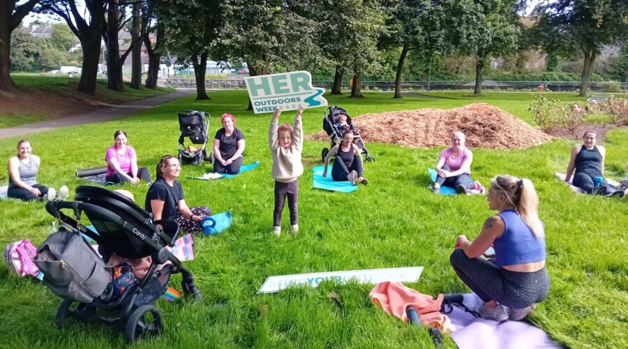 Moneymore Hub - Women sit on grass while a child in the centre stands tall with a sign for HER Outdoors week.