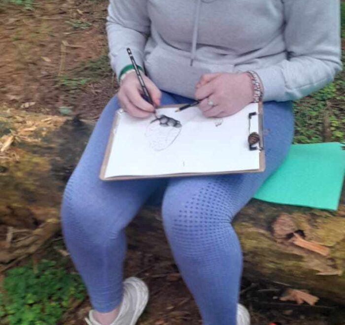 Girl draws on a clipboard while sitting on fallen tree trunk.