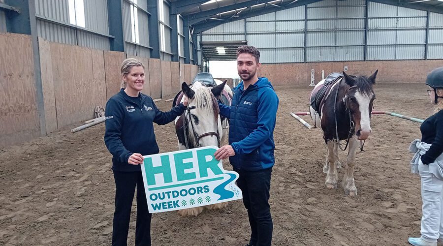 Man and woman pet horse while holding sign for Her Outdoors Week