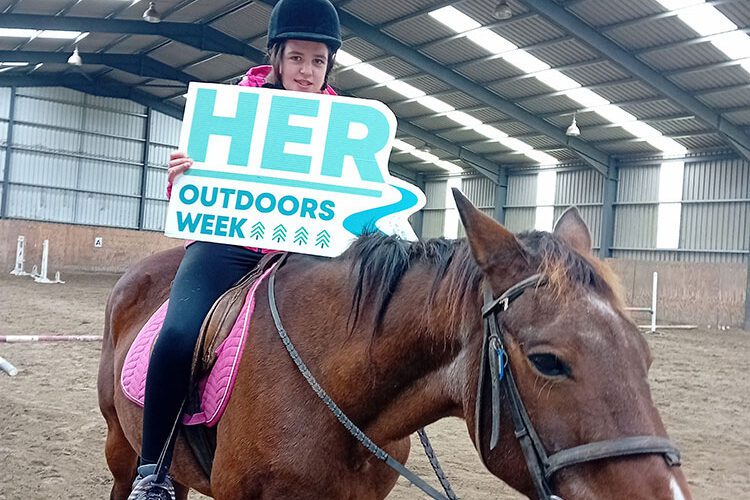 Young girl rides a brown horse and holds sign for Her Outdoors Week.
