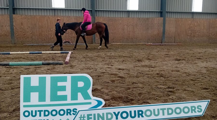 Sign for Her Outdoors Week with a young girl riding a horse in background.