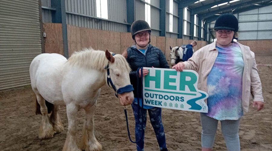 Two young girl hold sign for Her Outdoors Week next to white pony.
