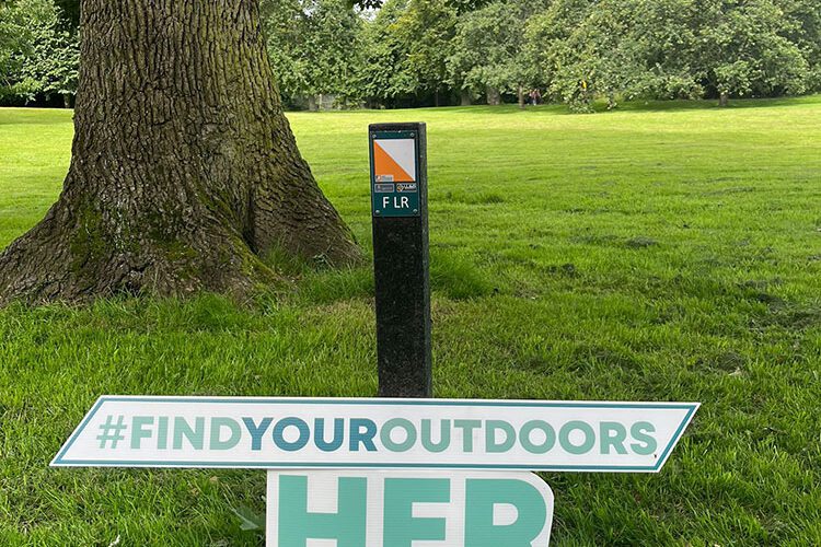 Sign for Her Outdoors Week rests against guide pole in park.