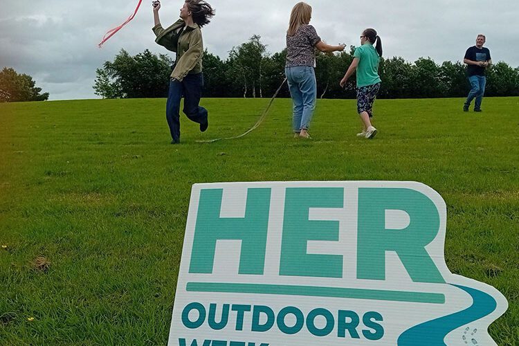 Sign for Her Outdoors Week with a family flying kites in the background.
