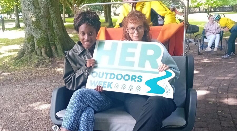 Two women sit holding a HER Outdoors sign.