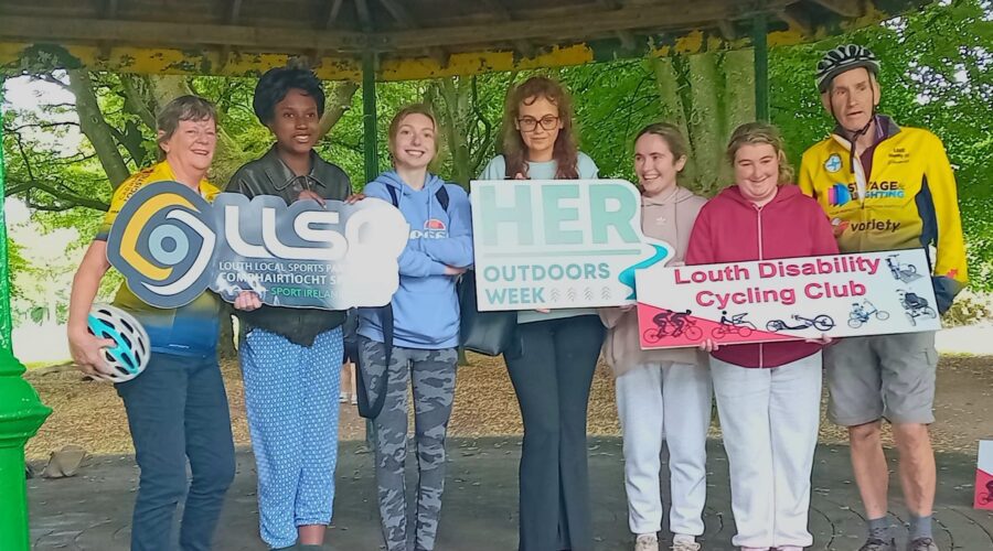 Group of people hold signage for LLSP, HER Outdoors week and Louth Disability Cycling Club under a gazebo.