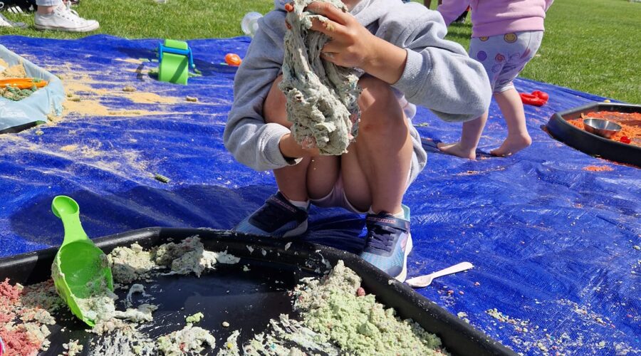 Young children play with sand on a mat on a pitch.