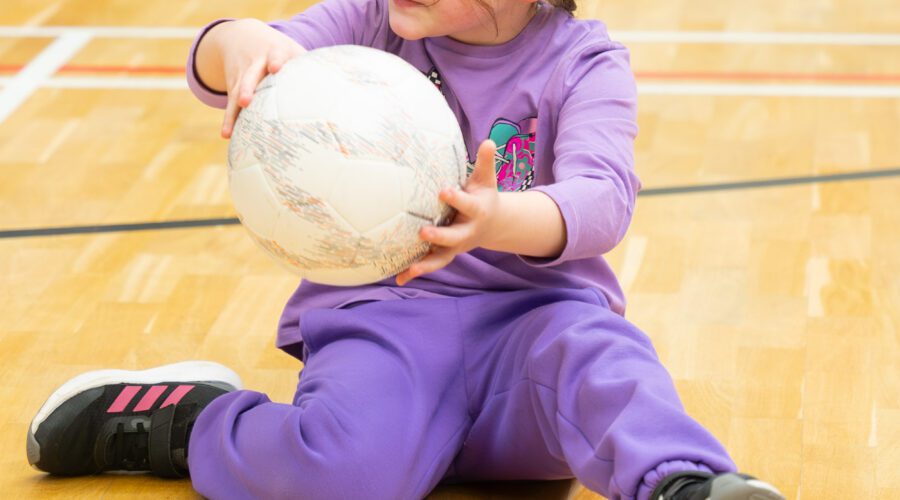 Young girl holds volleyball on floor of gym.