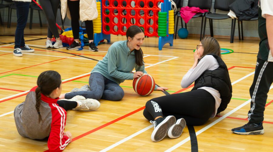 Three girls sit on floor of gym with basketball.