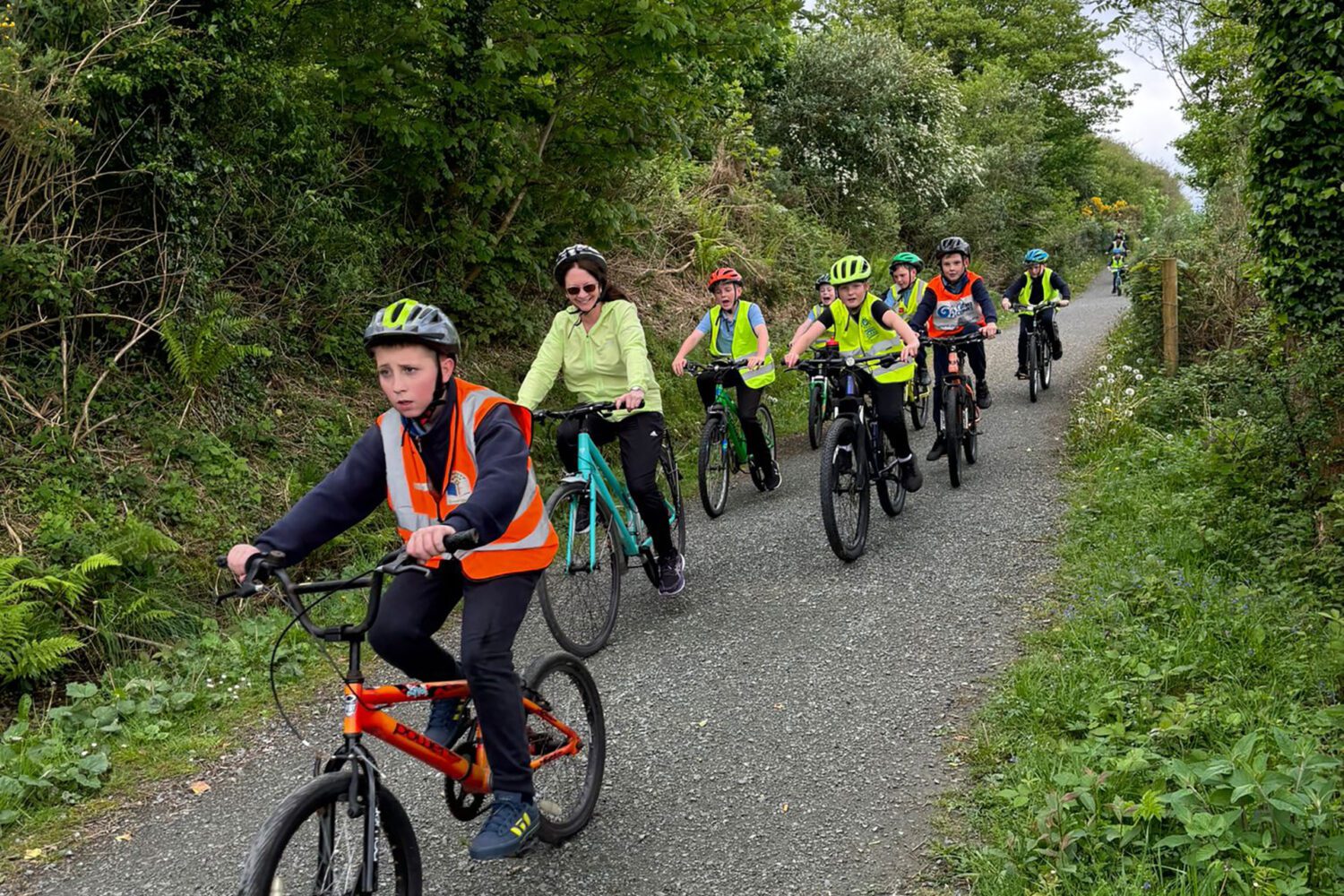 Young children cycle on a greenway wearing helmets and hi-vis jackets.